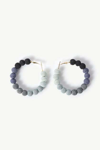 The Woven Ball Hoop earrings feature a circular design with gradient colored beads transitioning from dark blue to light gray, arranged on a white background, showcasing a subtle bohemian style.