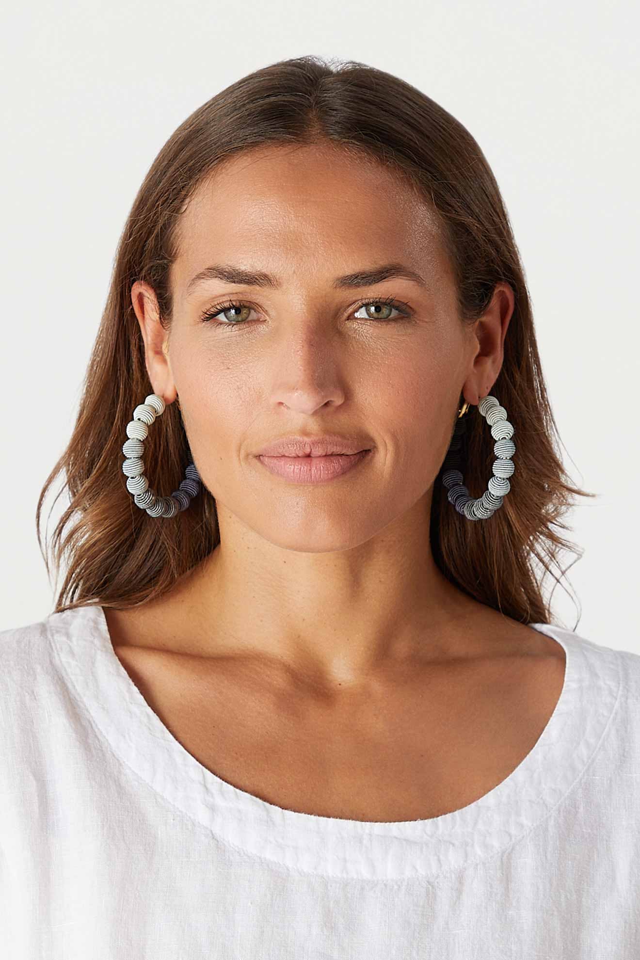 A person with long brown hair is wearing Woven Ball Hoops and a white shirt, looking directly at the camera against a plain background.