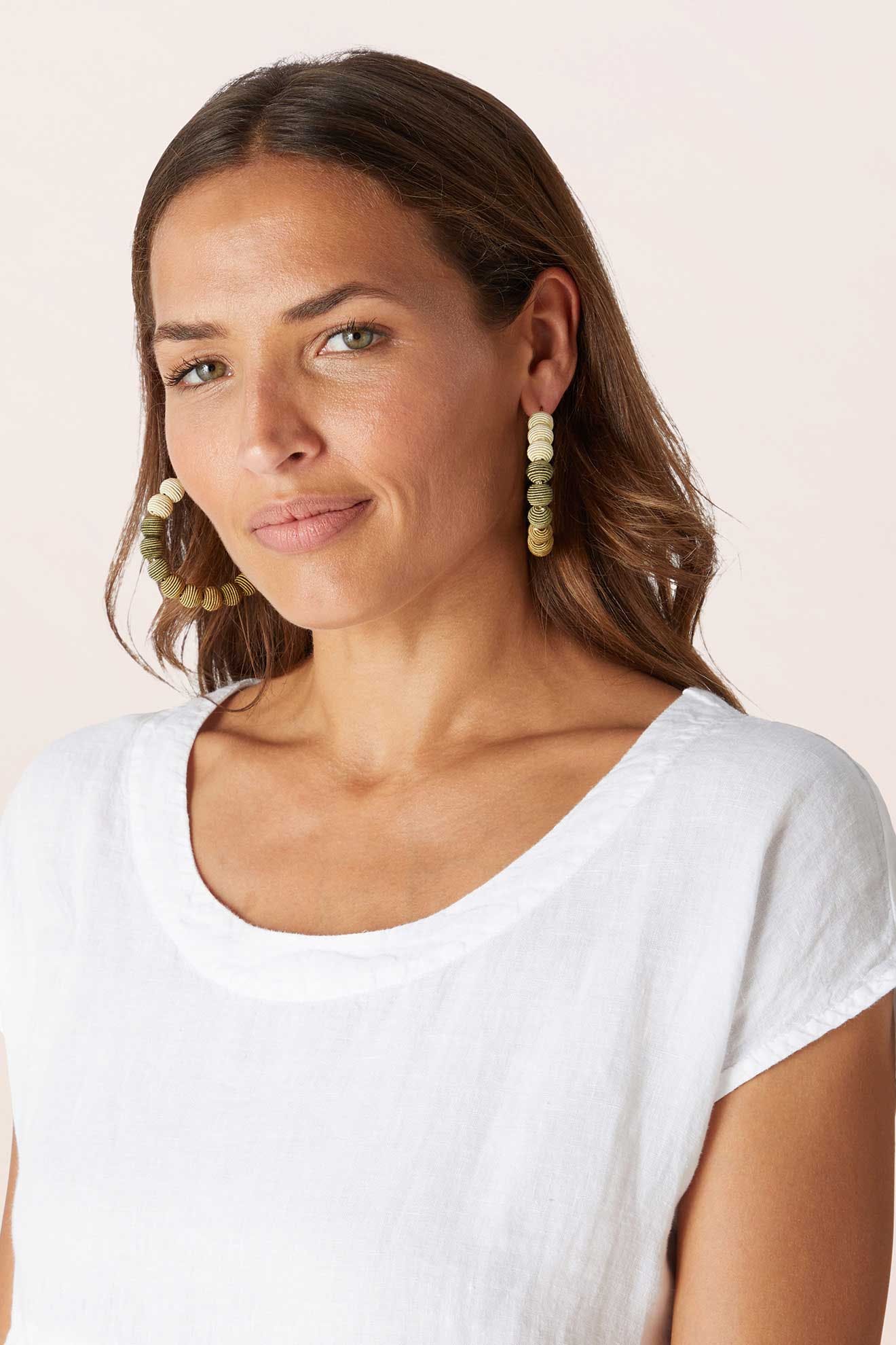 Woman with long brown hair, wearing a white top and Woven Ball Hoop earrings, faces forward with a neutral expression against a light background.