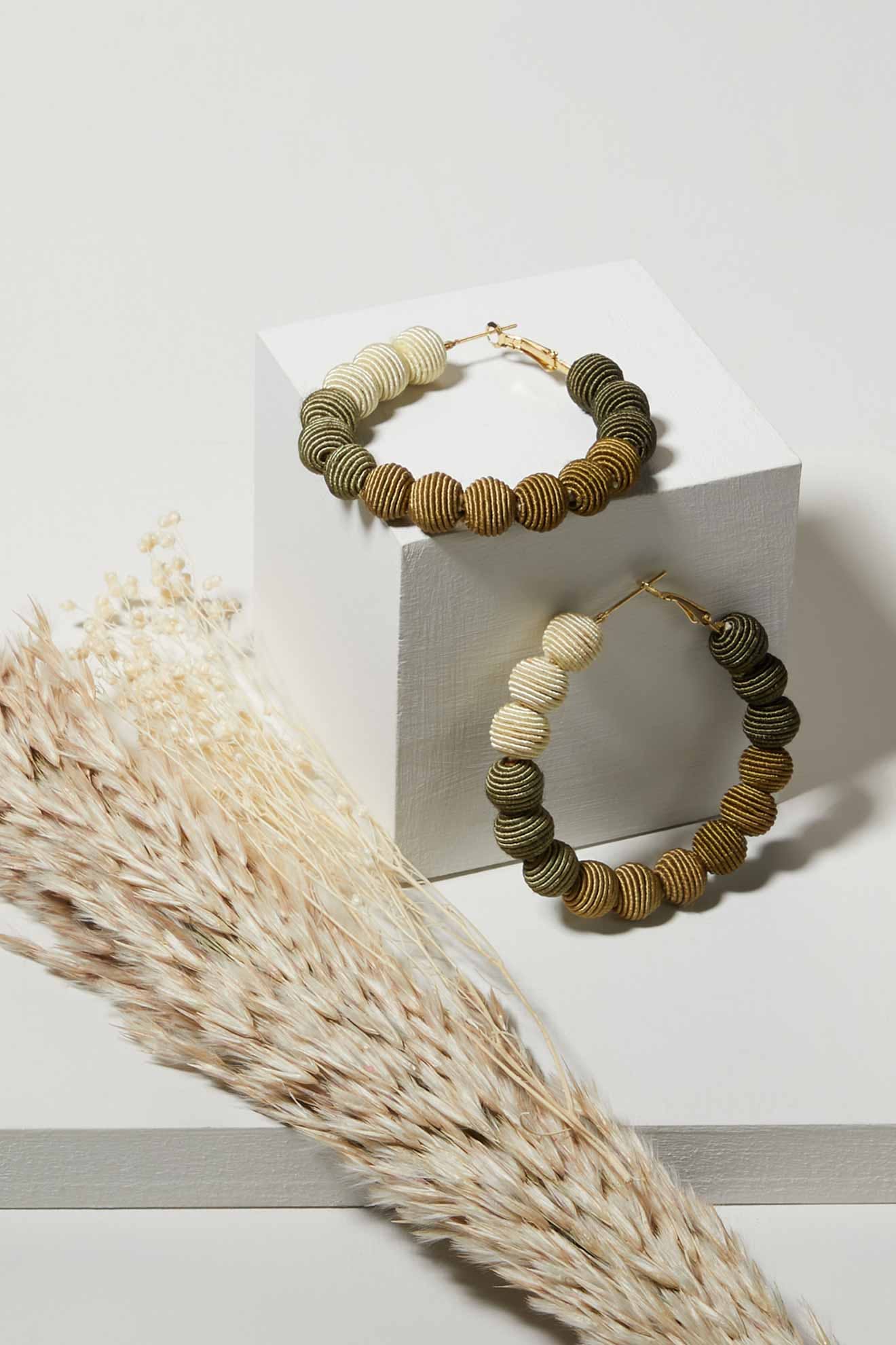 The Woven Ball Hoop earrings, featuring textured circular design in neutral hues, are showcased on a white block with dried pampas grass for a bohemian vibe.