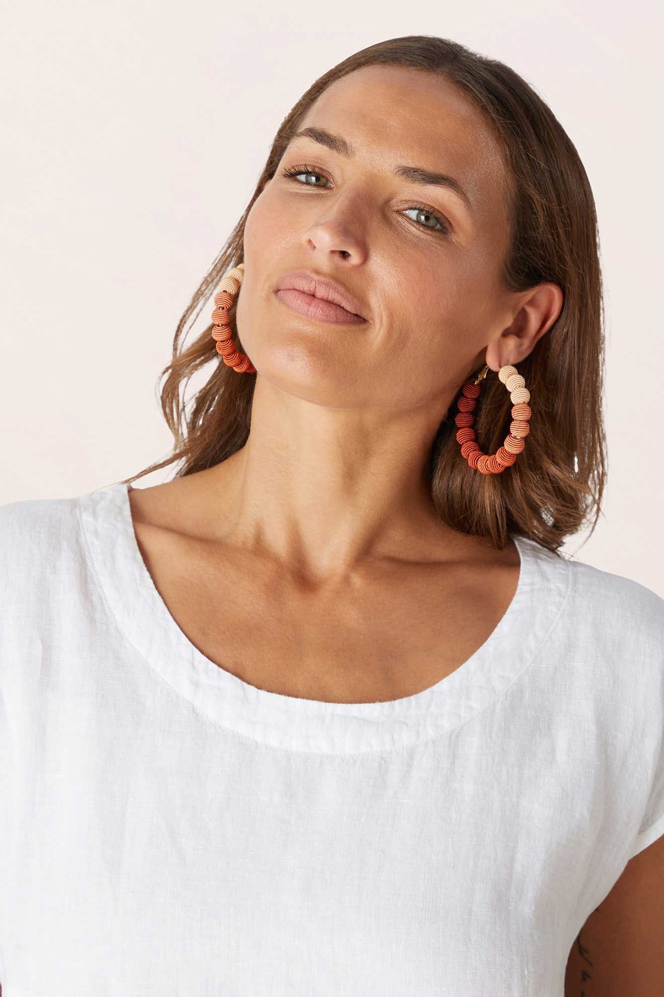 A woman with long brown hair wears a white linen top and Woven Ball Hoop earrings in bold orange and peach, looking confidently at the camera.