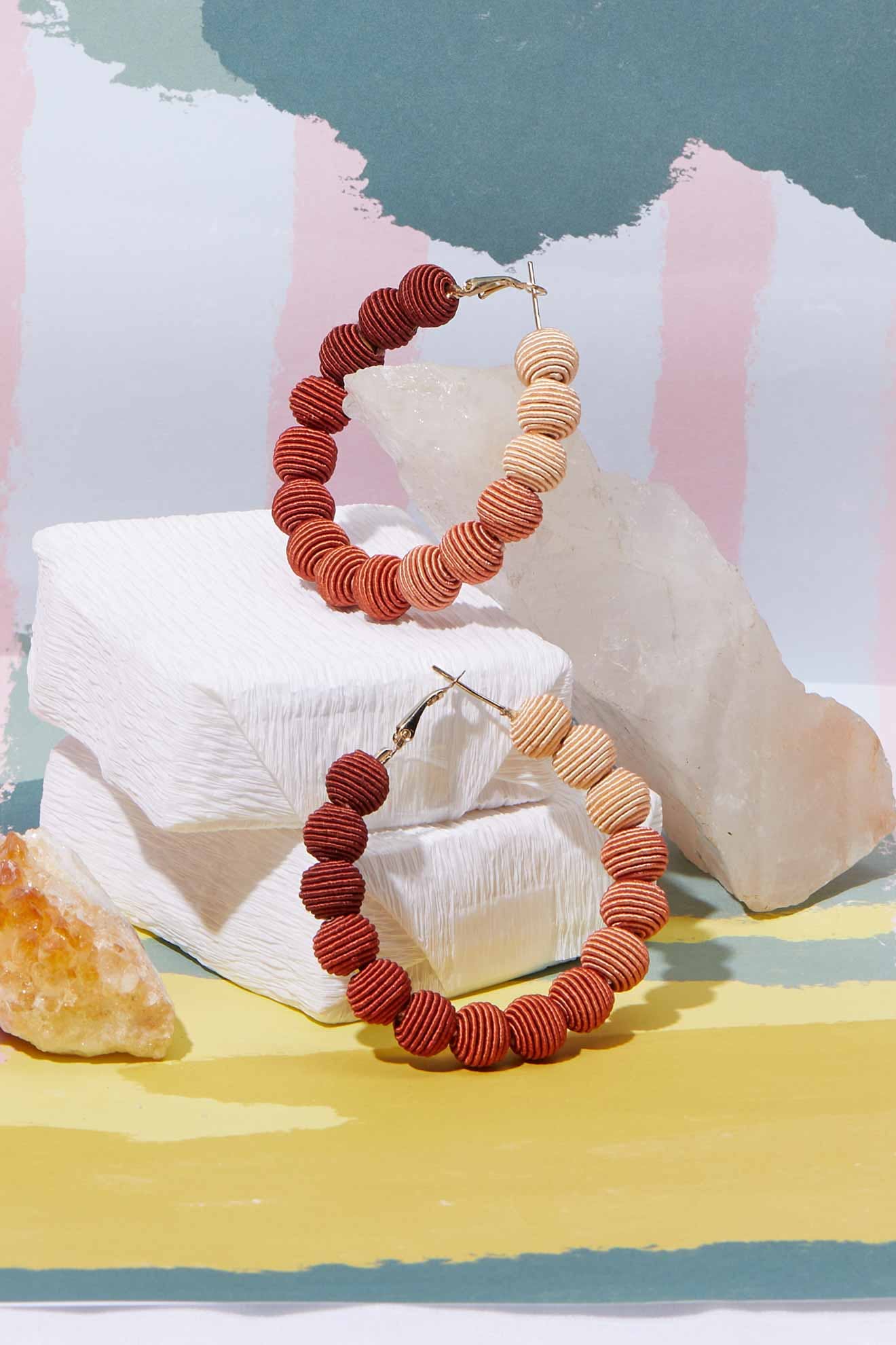 The Woven Ball Hoop earrings, featuring large bohemian-style hoops with alternating red and beige disc-shaped beads, are displayed on white textured blocks and rocks set against a pastel abstract backdrop.