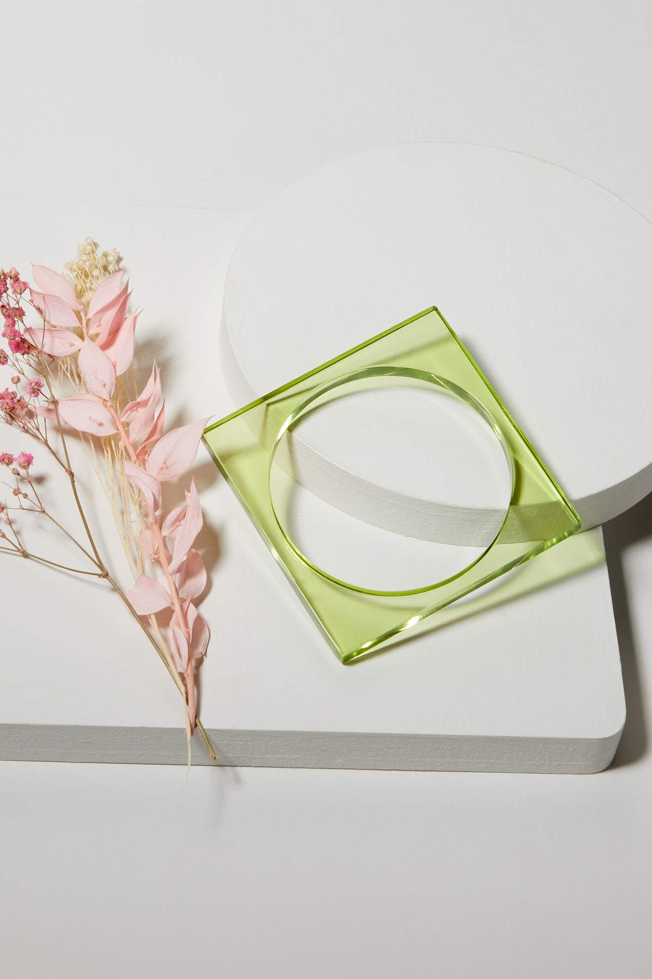 The Square Bangle, a transparent accessory, rests on white geometric platforms beside pink dried flowers—perfect for modern style collections.