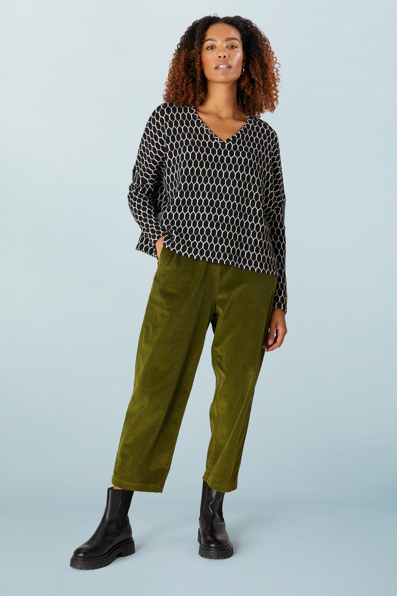 A person wears the Honeycomb Top, an oversized black and white patterned piece crafted with the flair of Italian artisans, paired with olive green pants and sleek black boots, all set against a plain background.
