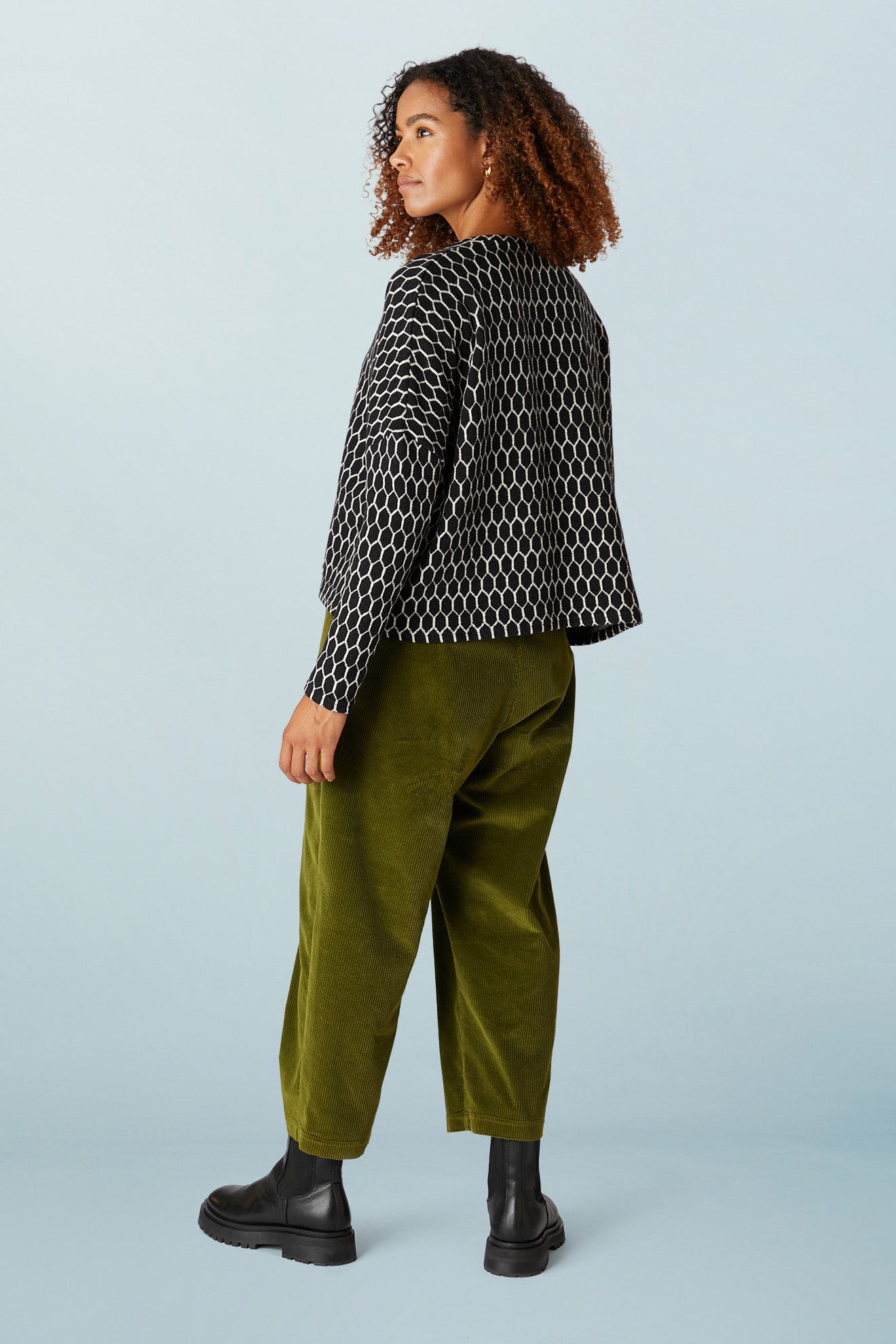 A person with curly hair is wearing an oversized black and white Honeycomb Top, olive green pants, and black boots, standing against a light blue background.