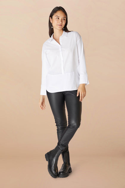 Sentence with Product Name: A woman stands wearing a white blouse and Black Vegan Leather Leggings, paired with black boots, against a light beige background.
