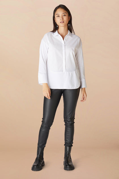 A woman stands confidently wearing a white blouse and **Ribbed Black Vegan Leather Leggings** paired with black boots, against a soft peach background.