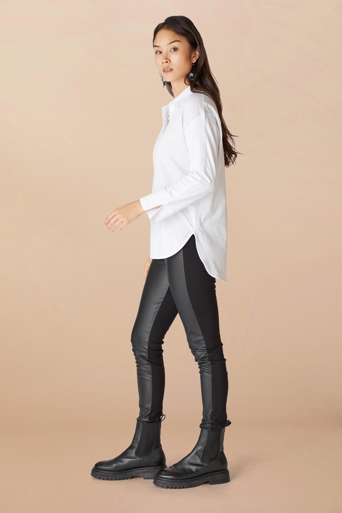Sentence with Product Name: A woman in a white shirt and Black Vegan Leather Leggings stands sideways, wearing black boots, against a pale background.