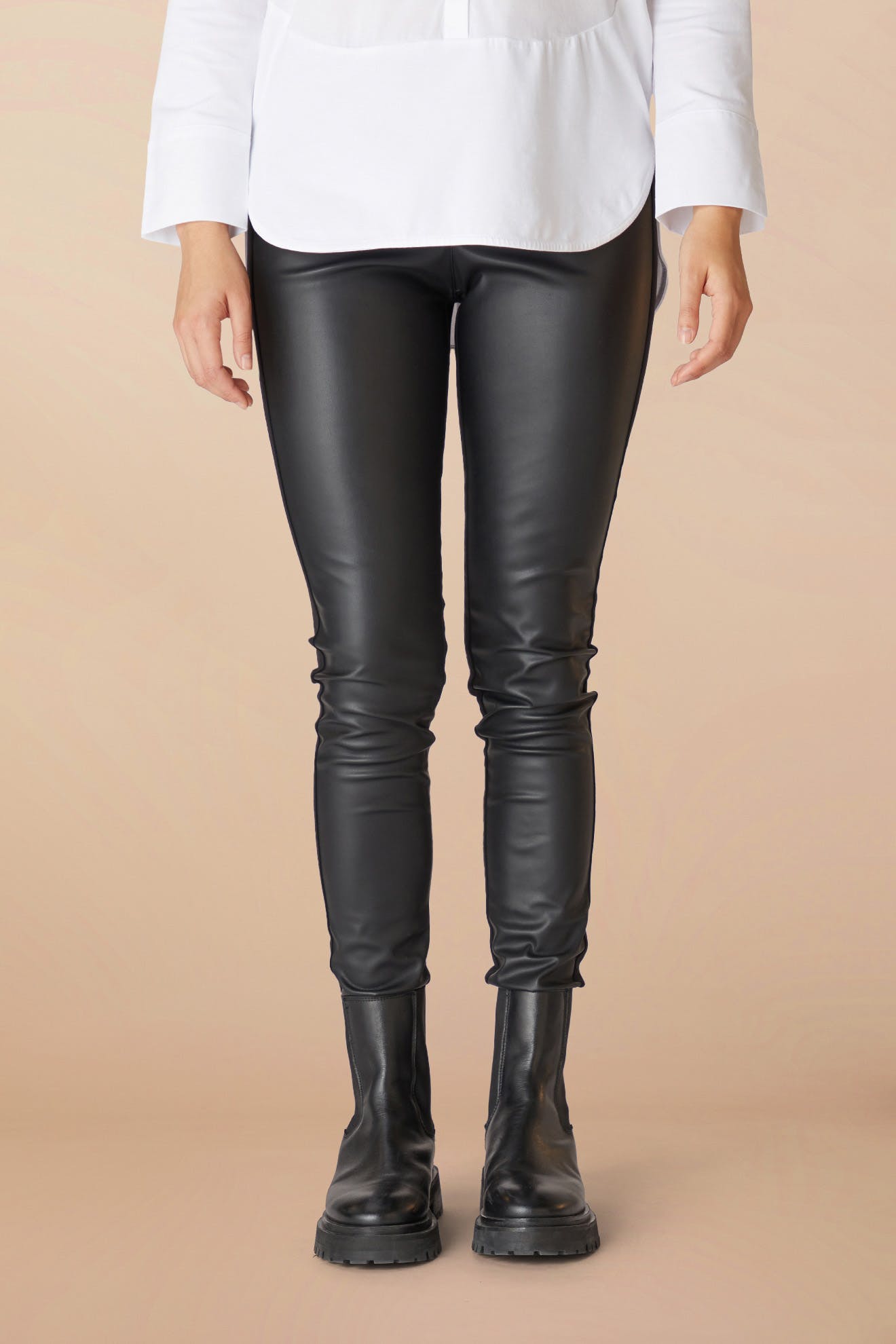 A person standing in Black Vegan Leather Leggings and boots, with a white shirt visible at the top, against a light beige background.