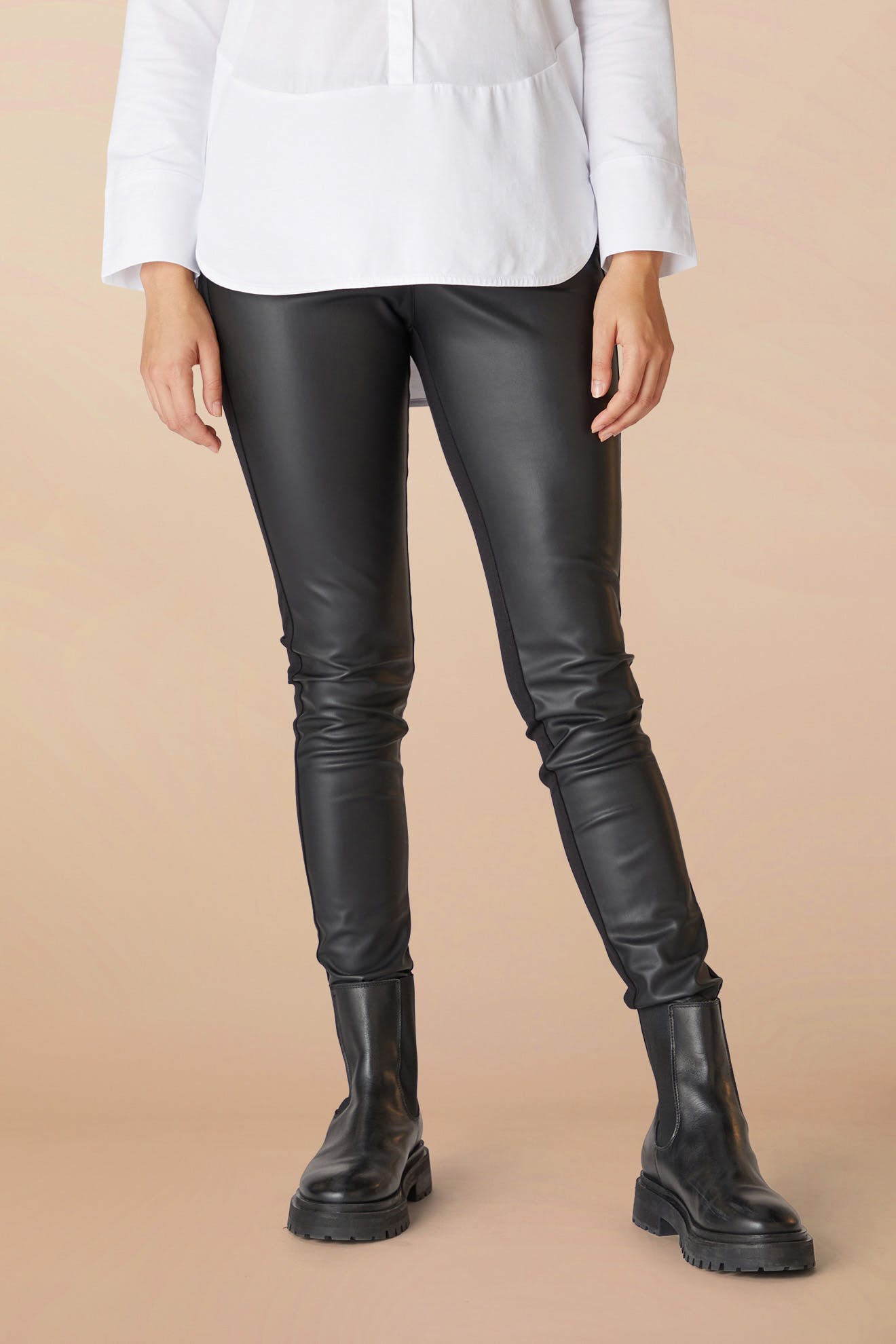 A woman stands wearing Black Maria Vegan Leather Leggings and black chunky boots, paired with a white shirt, against a soft peach background.