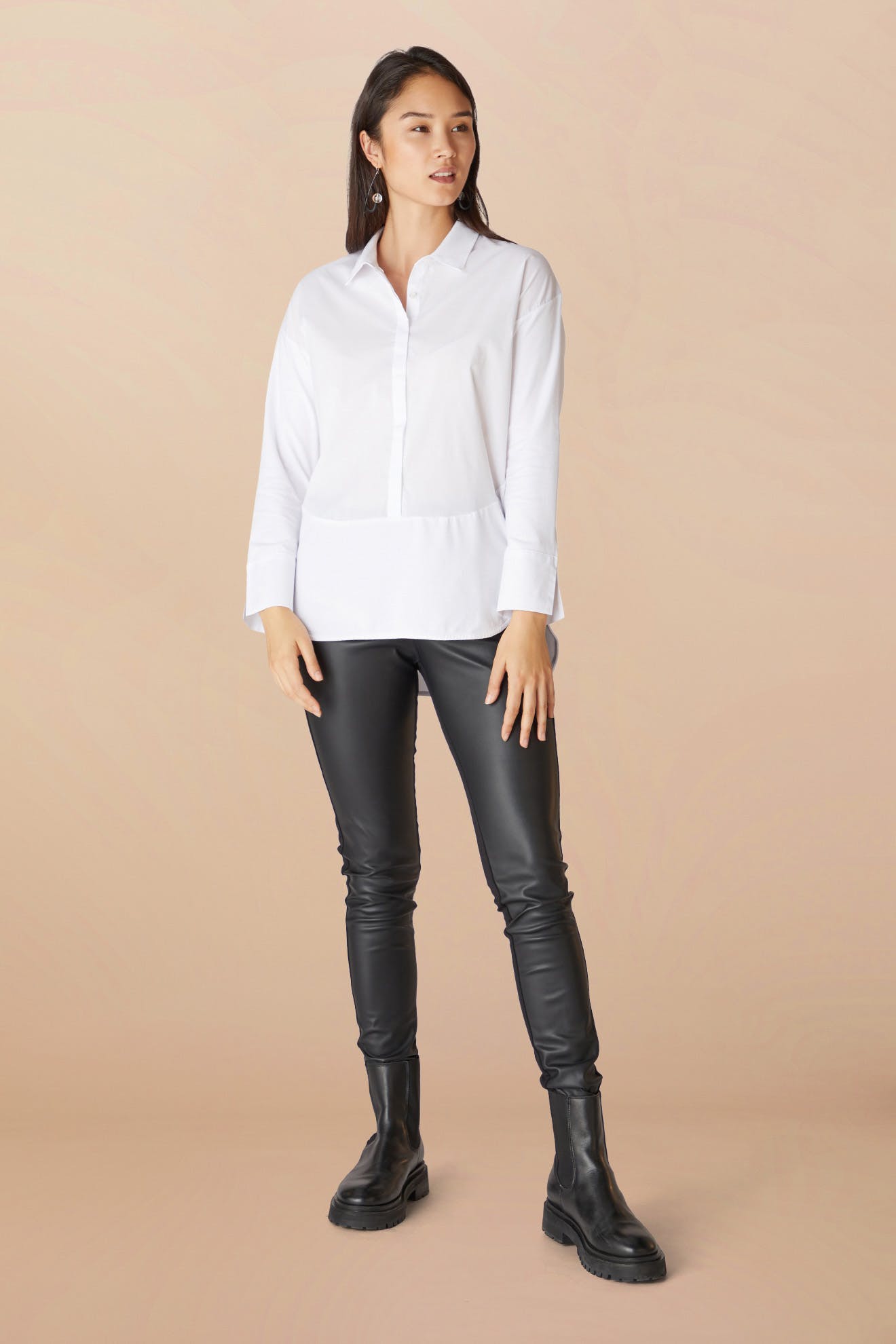Woman standing confidently in a white blouse and black vegan leather leggings, paired with black boots, against a soft peach background.