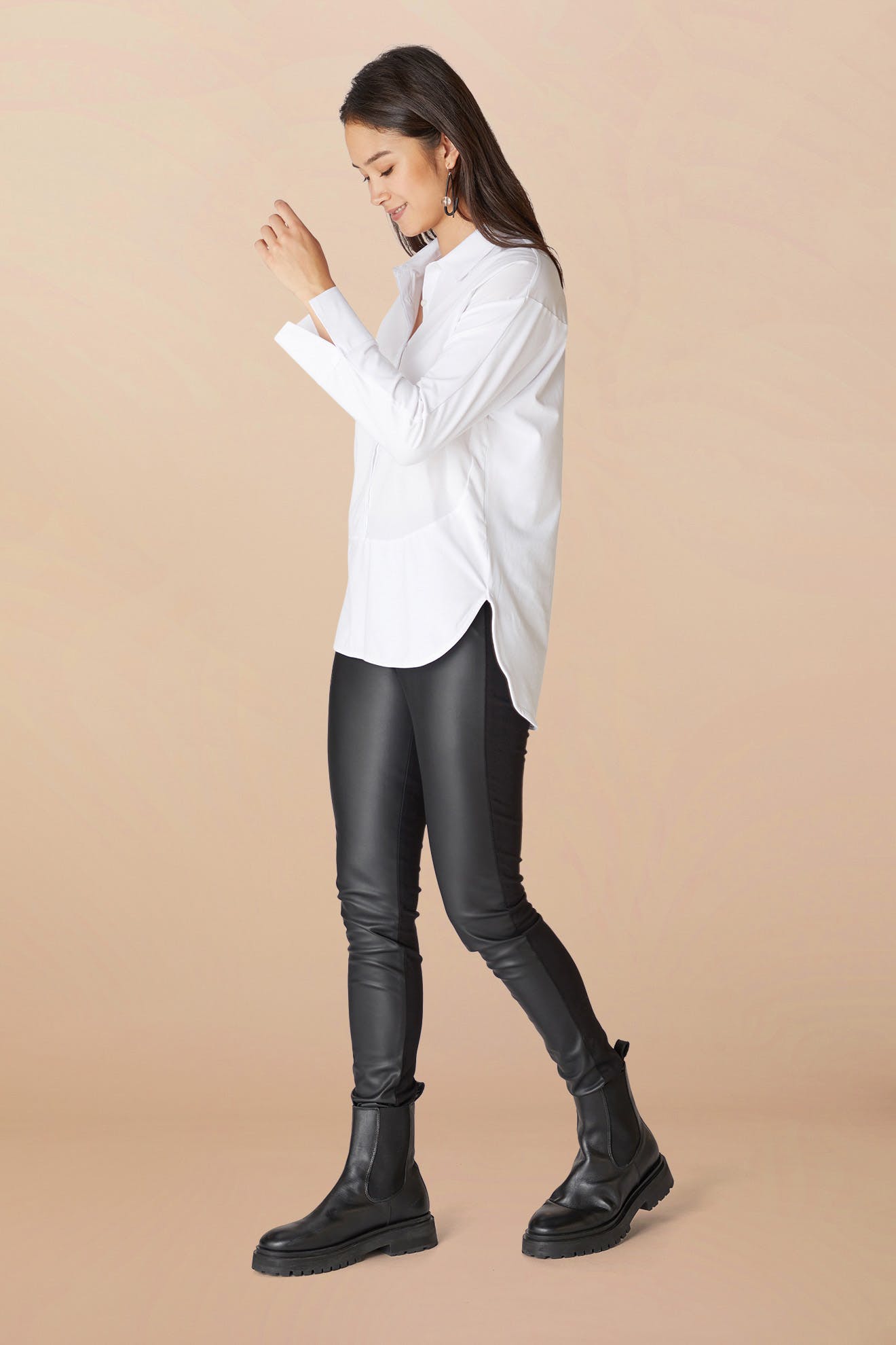 A woman in a white shirt and Black Vegan Leather Leggings is standing profile, smiling while looking at her wrist.