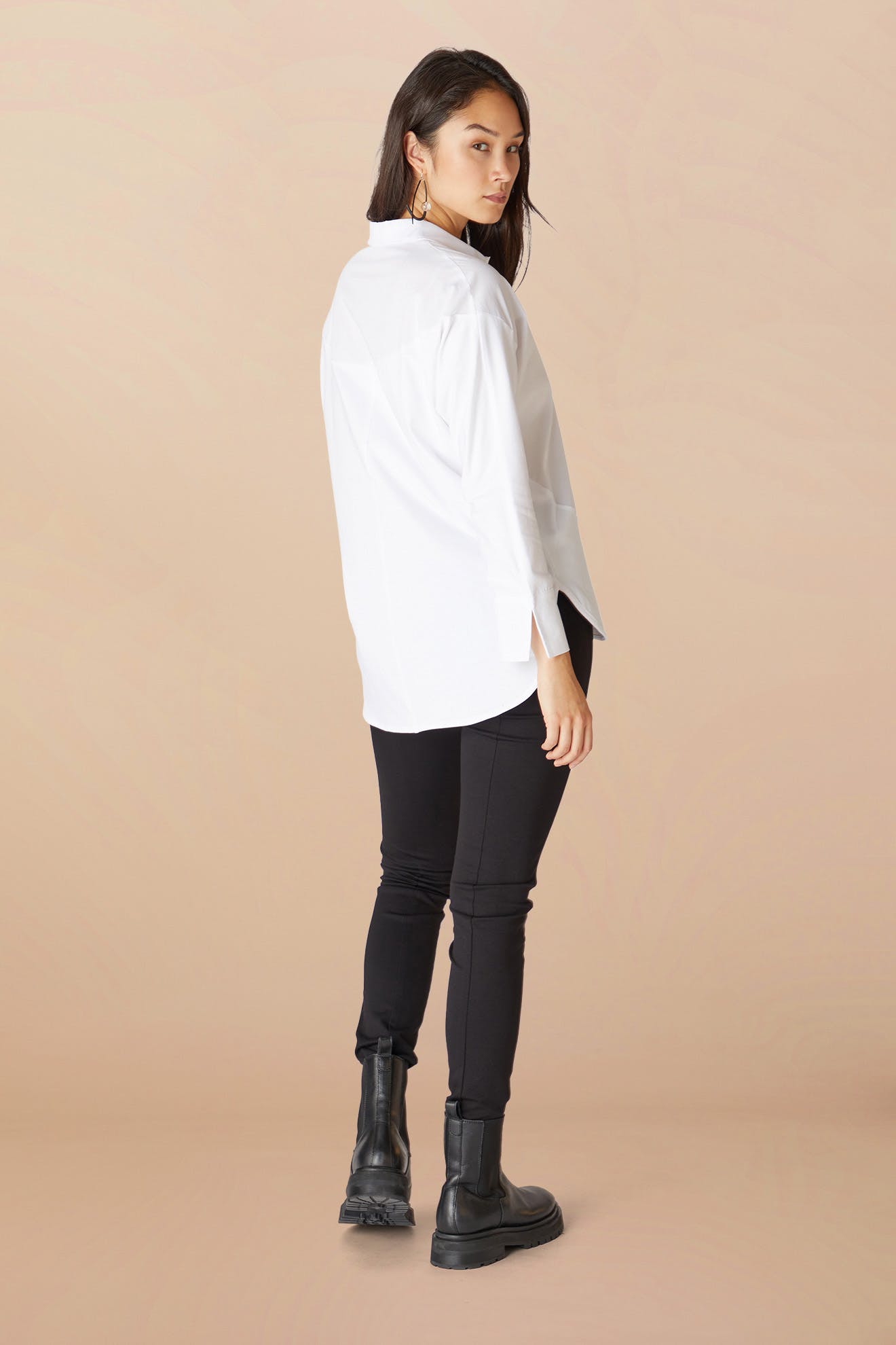 Sentence with Product Name: A woman stands in profile view, wearing a white oversized shirt and Black Vegan Leather Leggings, paired with black boots, against a pale background.