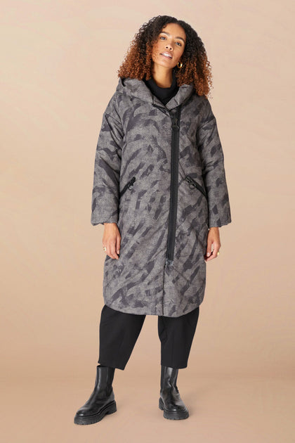 A woman in the Zip Print Coat—a long, patterned gray puffer with an asymmetrical zipper—black pants, and black boots stands against a beige background.