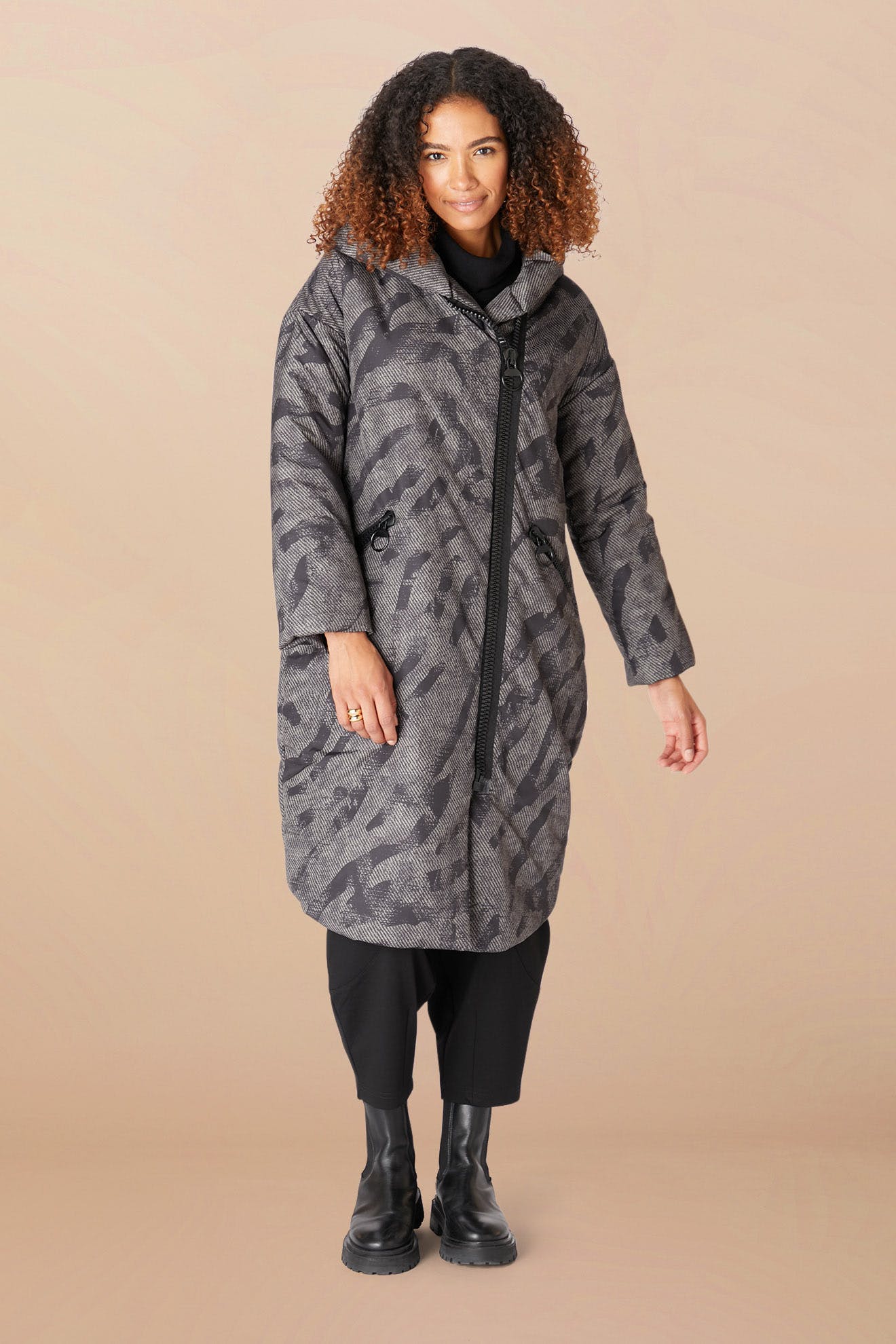 A woman faces forward in the Zip Print Coat—a long, patterned grey coat with a cozy hood and diagonal zipper—paired with black pants and boots, set against a beige background.