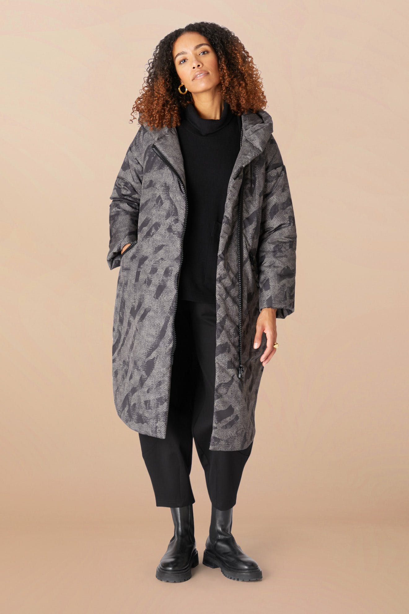 A woman stands against a beige background, wearing the Zip Print Coat—a gray patterned puffer with a cozy hood—paired with a black turtleneck, black pants, and black boots, her hands tucked in her coat pockets.
