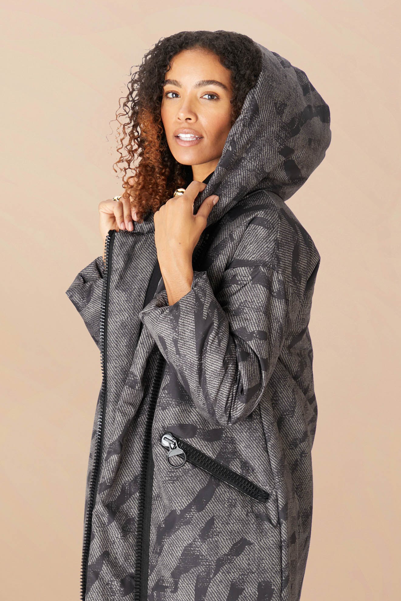 A woman wears the Zip Print Coat, a cozy black and gray camouflage hooded jacket, holding up the hood with both hands against a plain beige background.