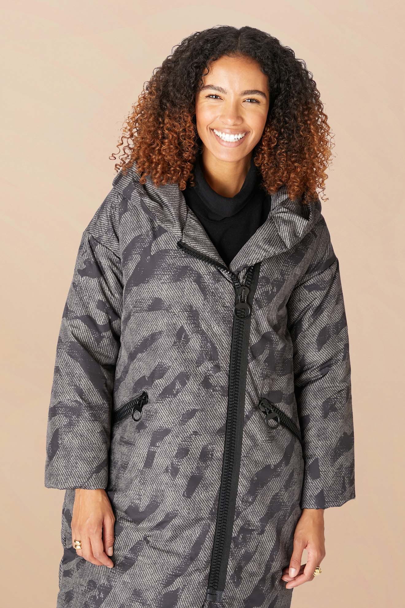 A woman with curly hair smiles while wearing the Zip Print Coat, featuring a long camouflage print and diagonal zipper, against a beige background.