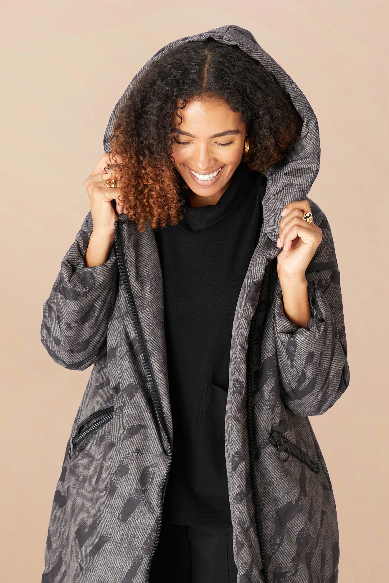 Woman smiles while holding up the cosy hood of the Zip Print Coat, a patterned gray puffer jacket, worn over a black top against a plain beige background.