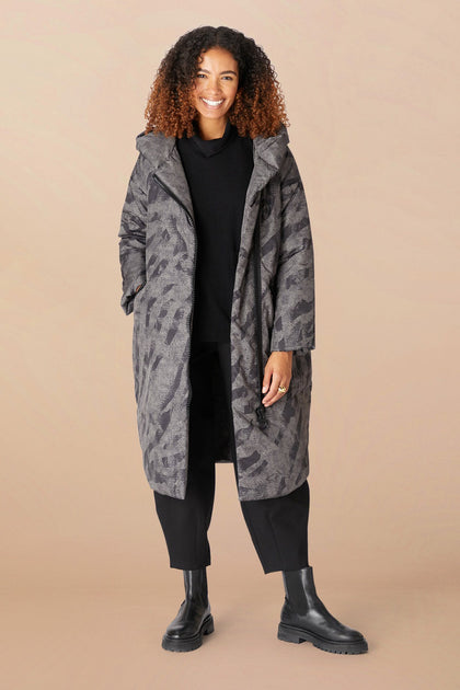 Woman stands wearing the Zip Print Coat, a long, patterned gray coat with a cozy hood, paired with a black sweater, black pants, and black boots against a plain beige background.