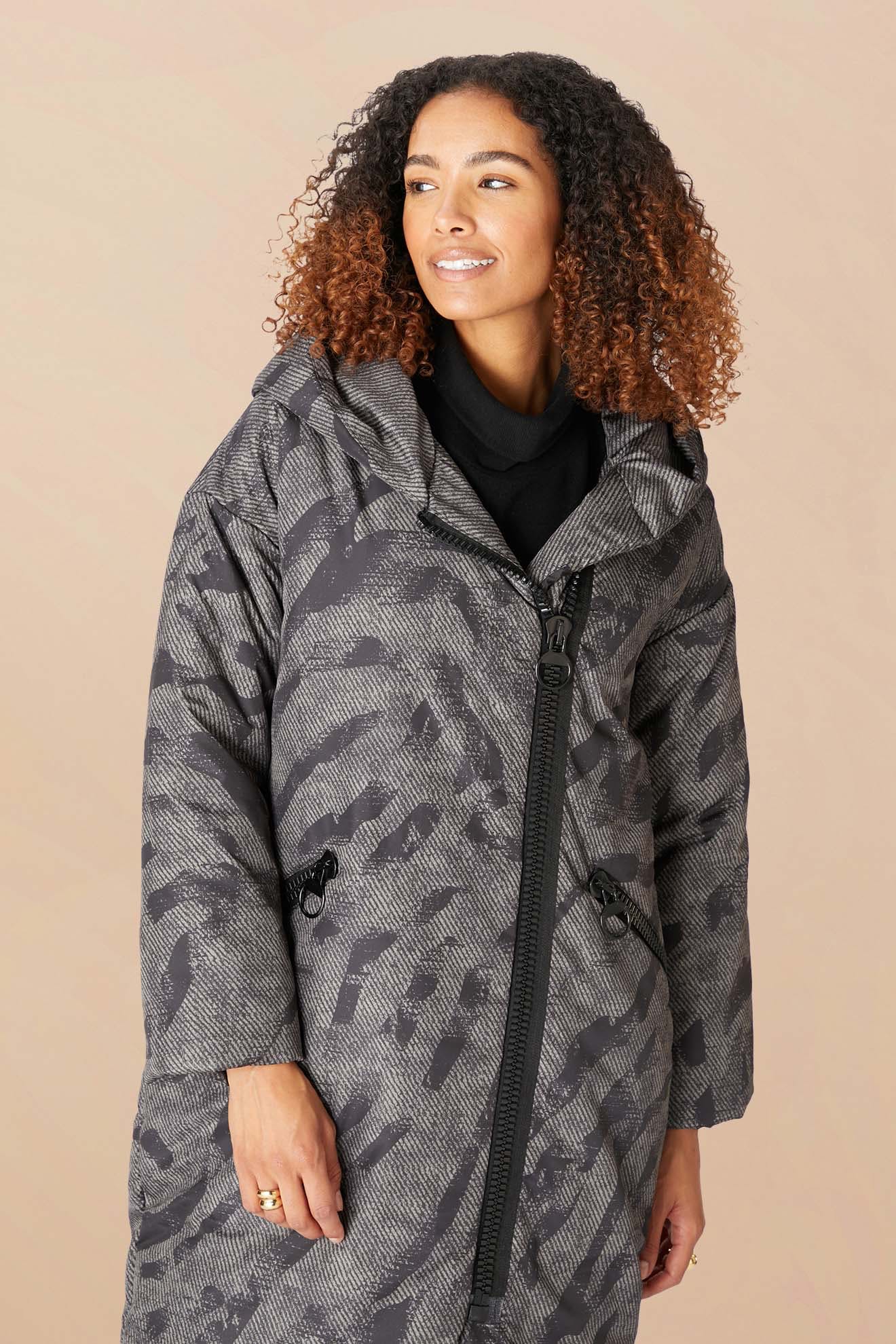 A woman with curly hair wears the Zip Print Coat, a patterned long gray puffer jacket with zipper and drawstrings, standing against a beige background.