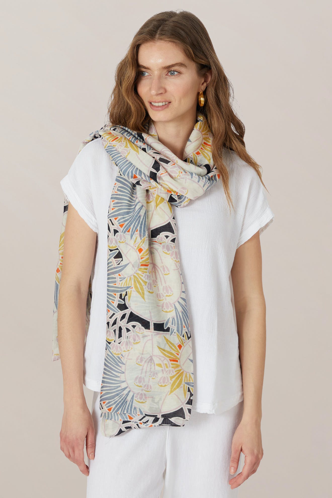 Woman with wavy brown hair, wearing a white modal viscose top and pants, accessorized with the Deco Daisy Scarf and gold hoop earrings, stands against a neutral background.