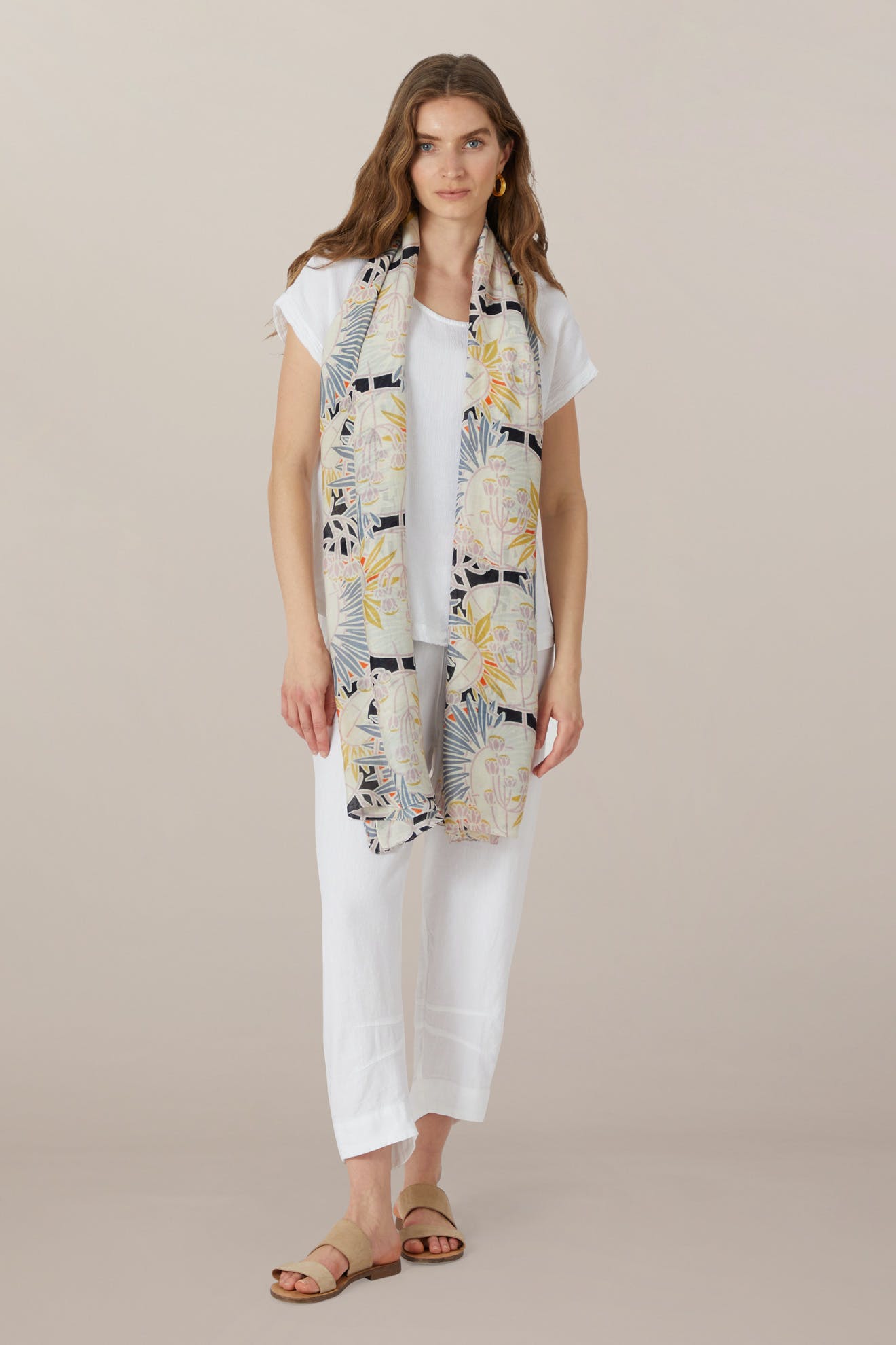 A woman stands facing forward, wearing a white short-sleeve top, white pants, tan sandals, and the Deco Daisy Scarf in soft modal viscose with a daisy print draped around her neck.