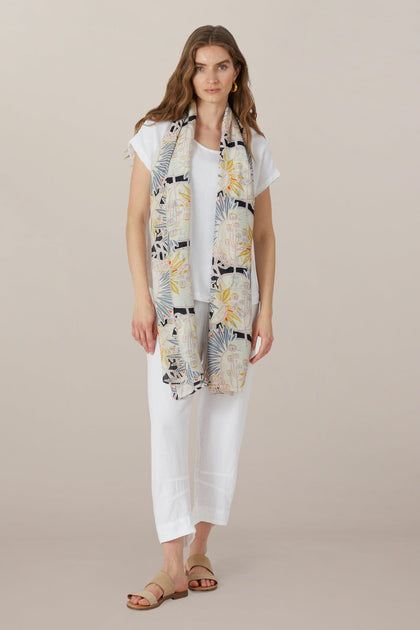 A woman stands facing forward, wearing a white short-sleeve top, white pants, tan sandals, and the Deco Daisy Scarf in soft modal viscose with a daisy print draped around her neck.
