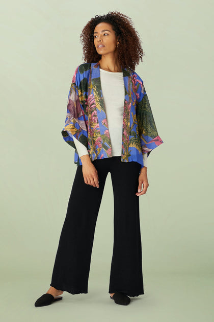 A person wearing the Kew Magnolia Kimono, crafted from lightweight modal viscose, stands gracefully against a light green background. It is paired with a white top and black wide-leg pants—creating a stunning ensemble reminiscent of the Kew Gardens collaboration.