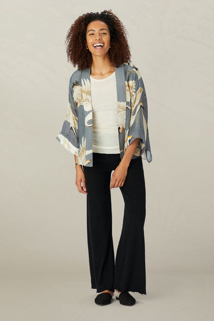 A person wearing the Stork Kimono from the Kew Gardens collaboration, paired with a white top and black pants, is smiling against a plain background.
