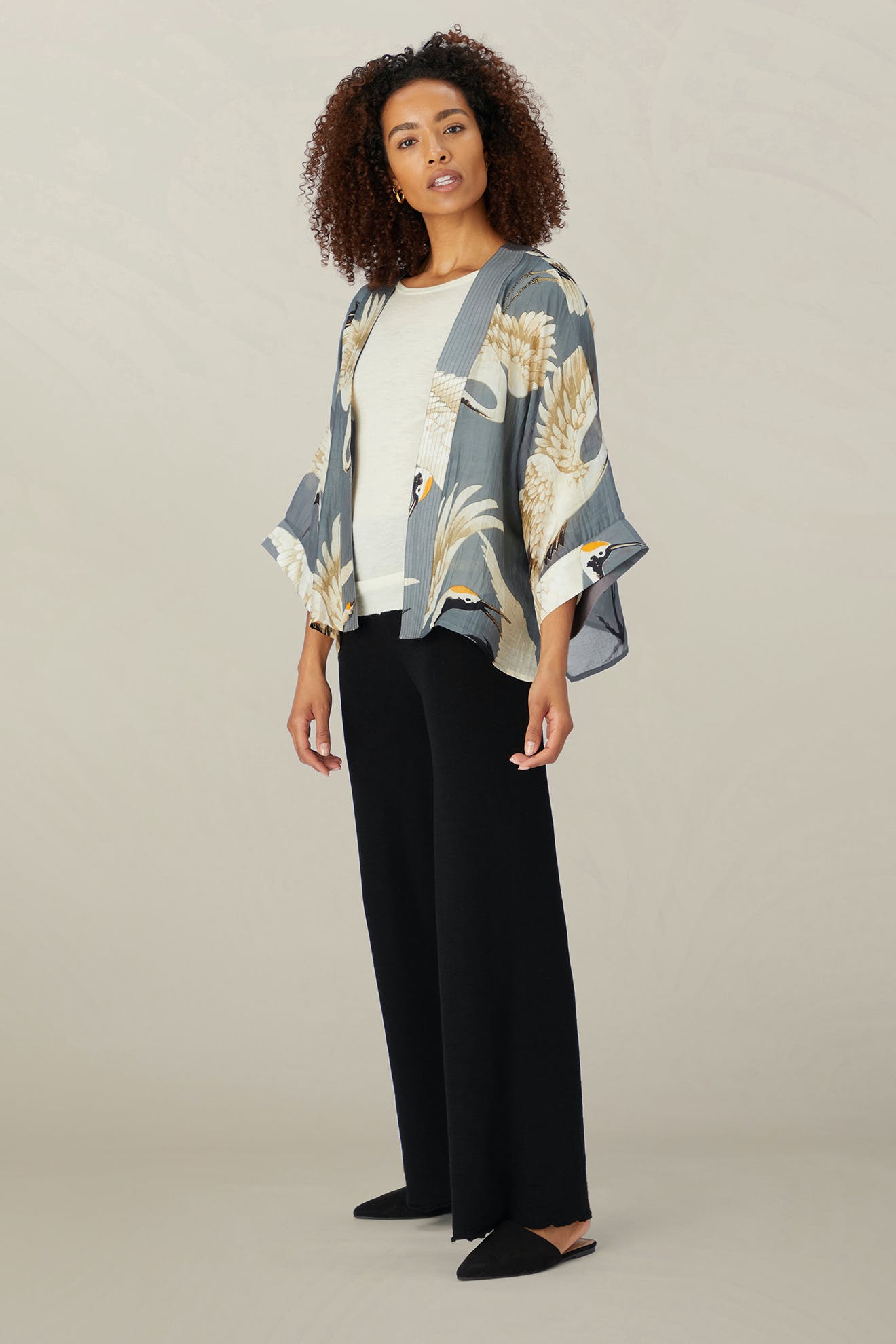 A woman stands gracefully against a neutral backdrop, draped in the Stork Kimono from the Kew Gardens collaboration. Her ensemble includes a crisp white shirt, black pants, and shoes, accentuating the kimono's vibrant floral design.