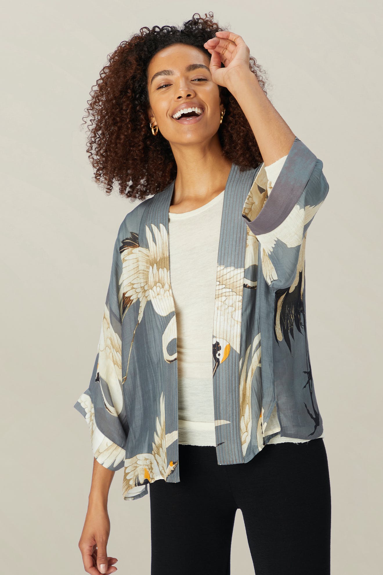A person with curly hair smiles, wearing the Stork Kimono from the Kew Gardens collaboration over a white top and black pants, against a plain background.