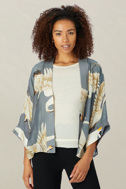 A person with curly hair wears the Stork Kimono, a printed jacket from the Kew Gardens collaboration, over a white shirt and pairs it with black pants. They stand against a neutral background, highlighting the fusion of style and nature in this lightweight modal viscose creation.