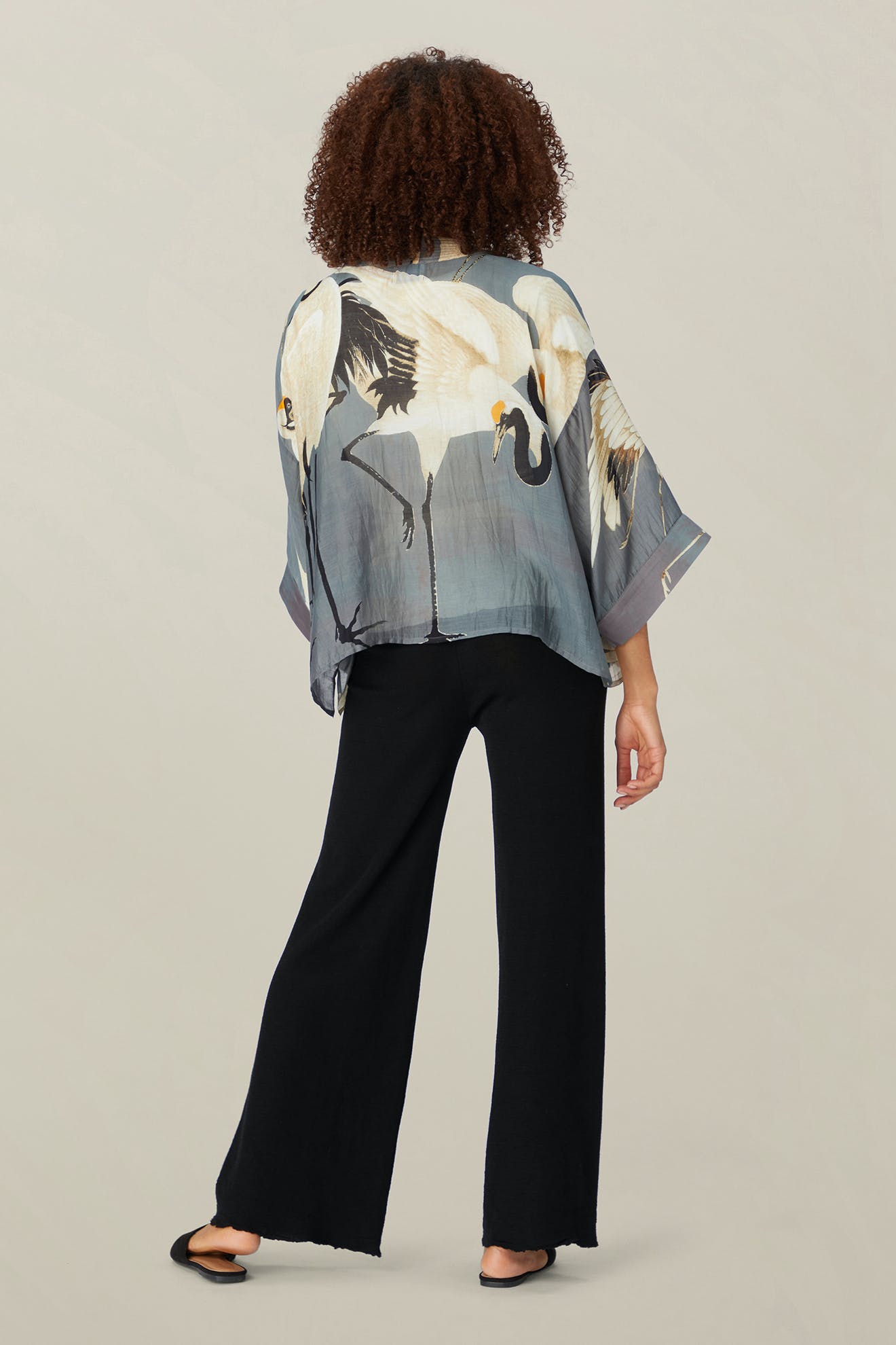 The individual with curly hair is dressed in the Stork Kimono, a lightweight modal viscose top styled like a kimono and displaying a crane design, complemented by black pants. This printed piece is part of the Kew Gardens collaboration and is captured from the back against a plain background.