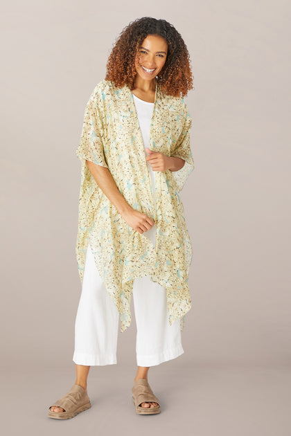 A woman smiles, wearing the Blossom Blue Throwover by One Hundred Stars over a white top and pants with beige sandals, posed against a neutral background.