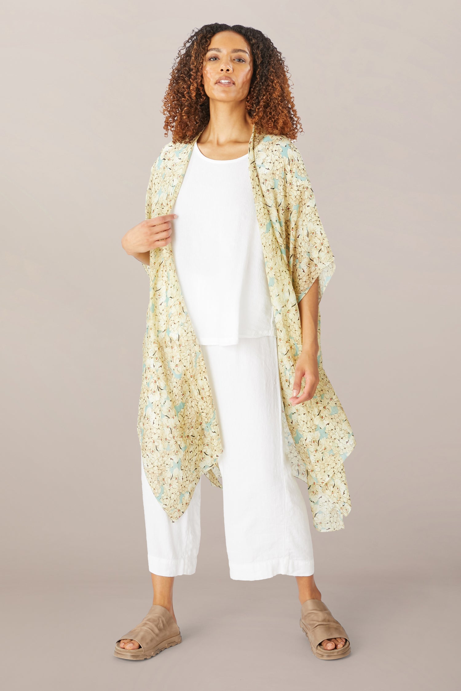 Person standing against a plain background, wearing a white outfit with the Blossom Blue Throwover and beige sandals.