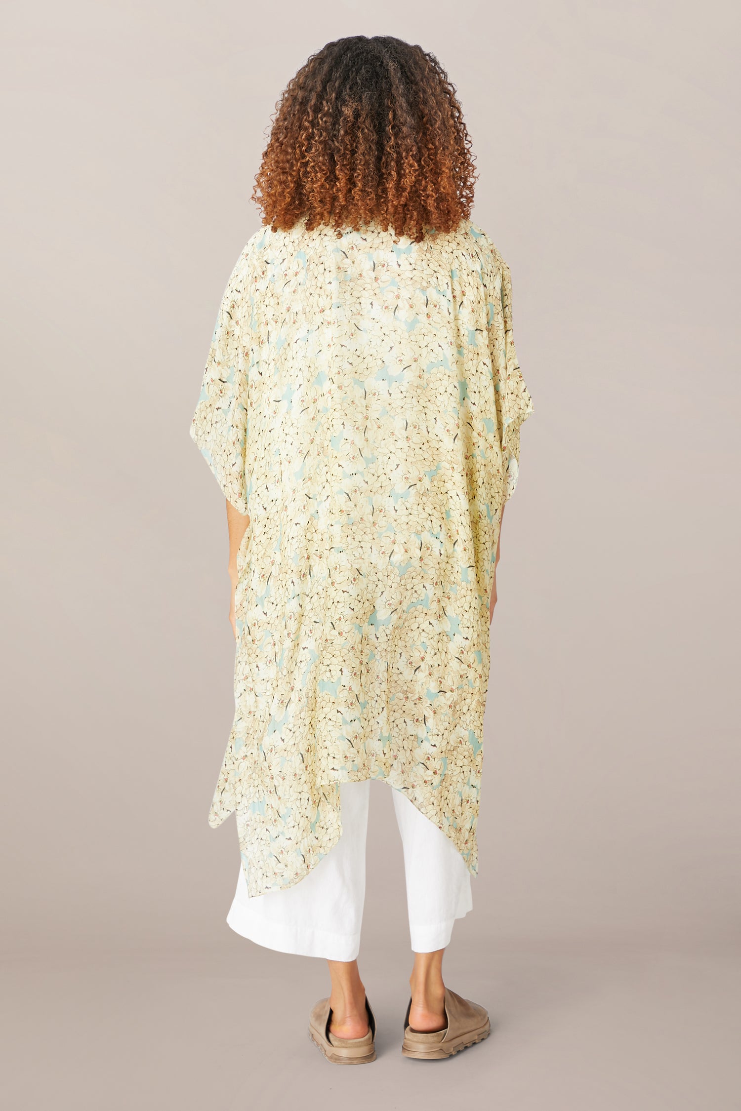 Person standing with their back to the camera, wearing a Blossom Blue Throwover, white pants, and beige shoes against a neutral background.