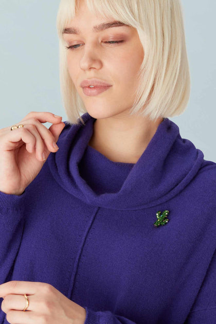 Person with short blonde hair in a purple sweater, wearing the Cactus Brooch, eyes closed, hand near their face.