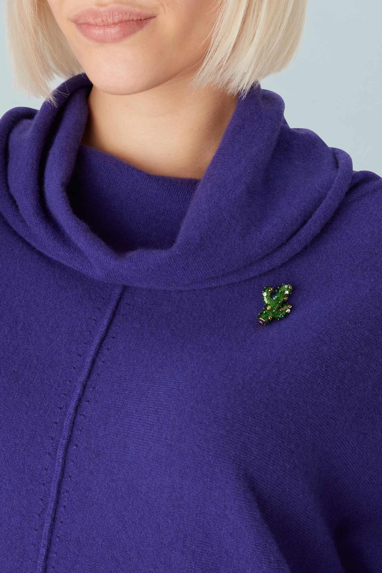A person with short blond hair wears a purple cowl-neck top with the Cactus Brooch—an intricate green jeweled metalwork piece—pinned by the shoulder.