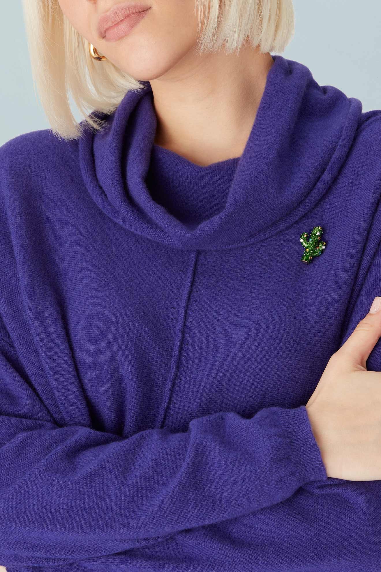 A woman with blonde hair, arms crossed, wears a purple cowl-neck sweater adorned with the Cactus Brooch against a light background.