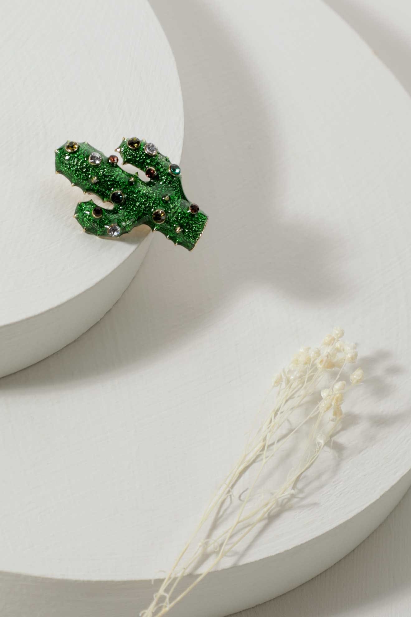 Cactus Brooch with green glitter and vibrant embellishments is displayed on a white circular platform next to dried white flowers.