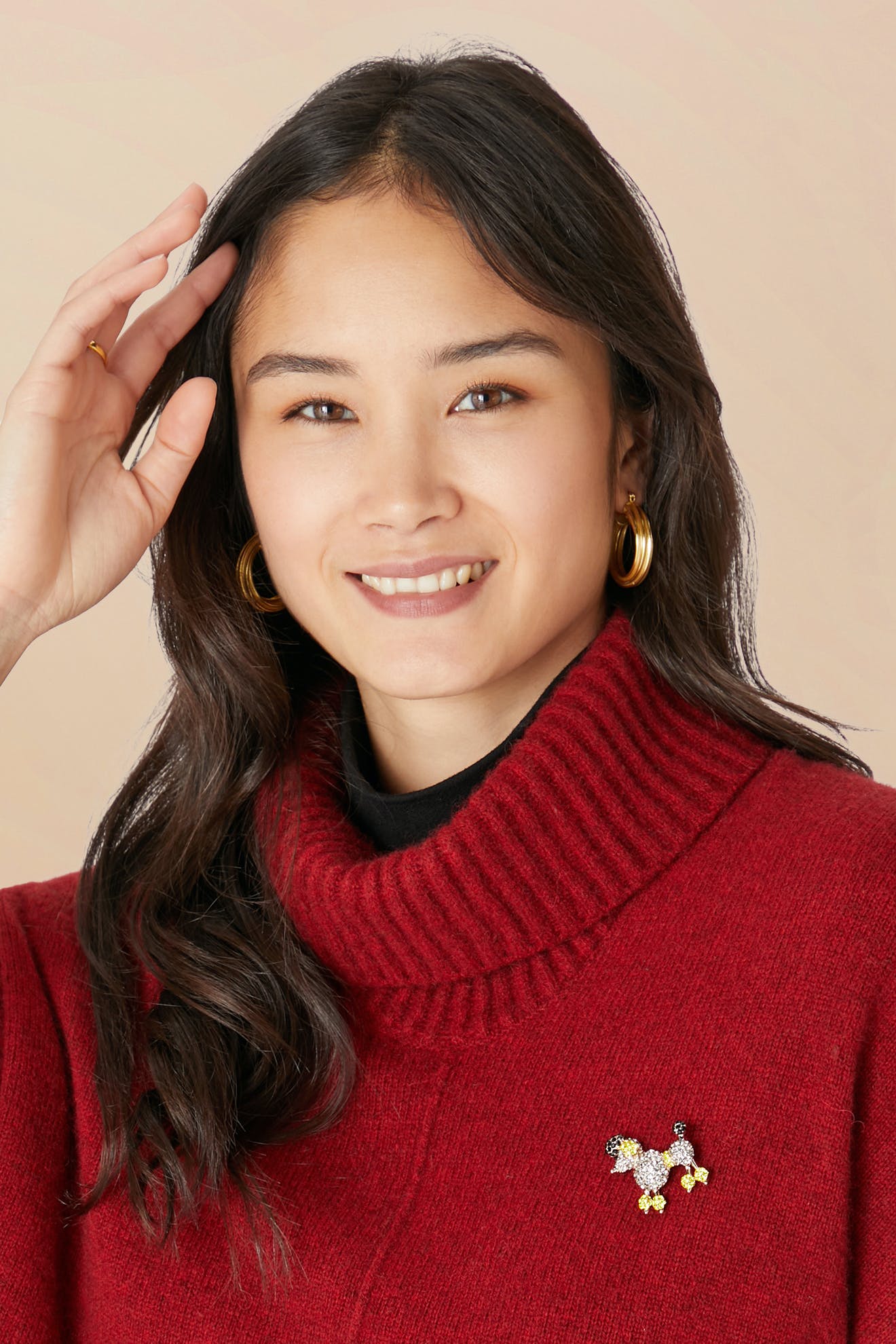Smiling person with long dark hair wears a red turtleneck, gold hoop earrings, and whimsical jewelry—a Poodle Brooch—raising one hand near their head.
