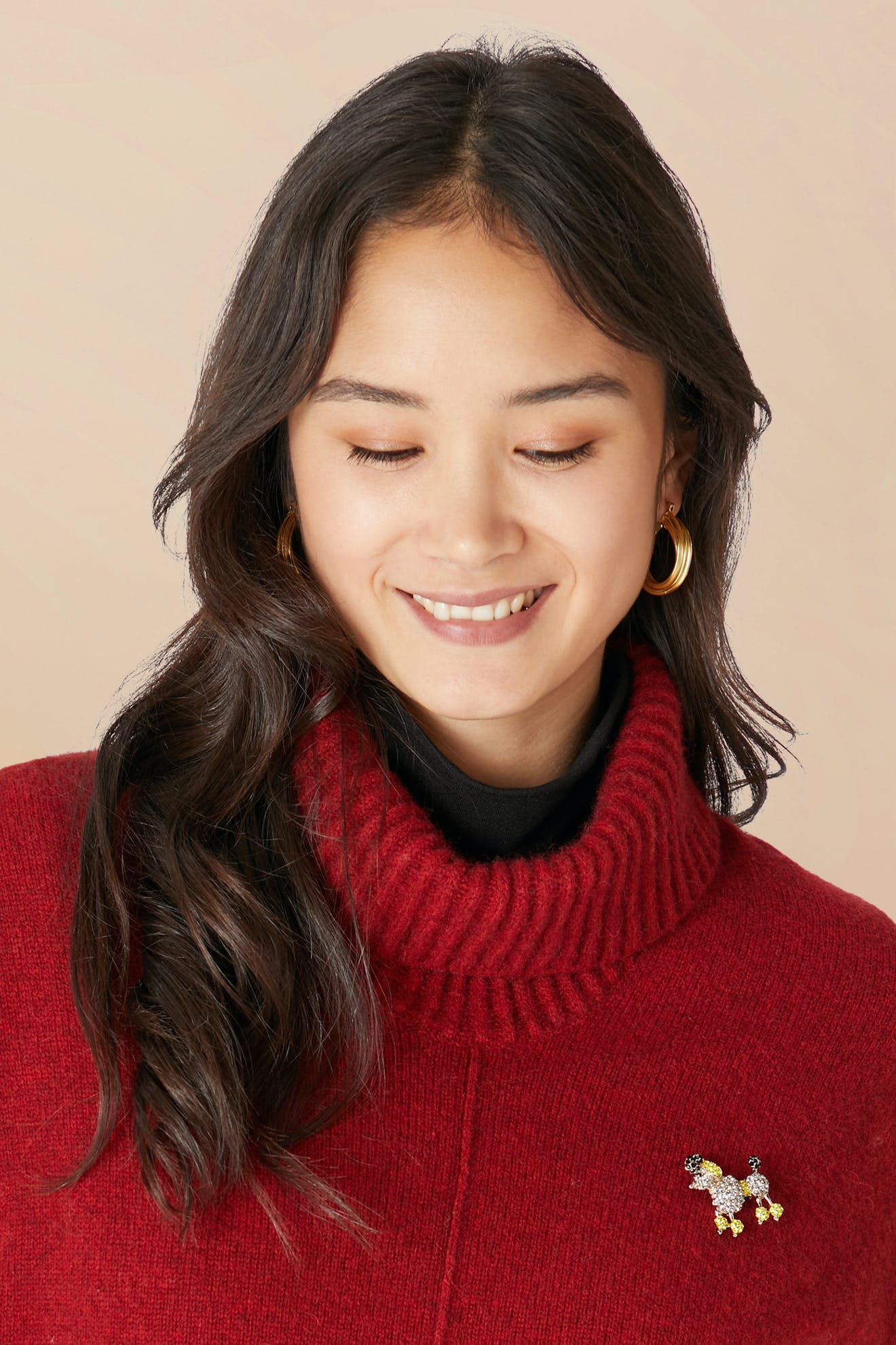 Woman with long dark hair in a red turtleneck and gold hoop earrings smiles while wearing the whimsical Poodle Brooch.