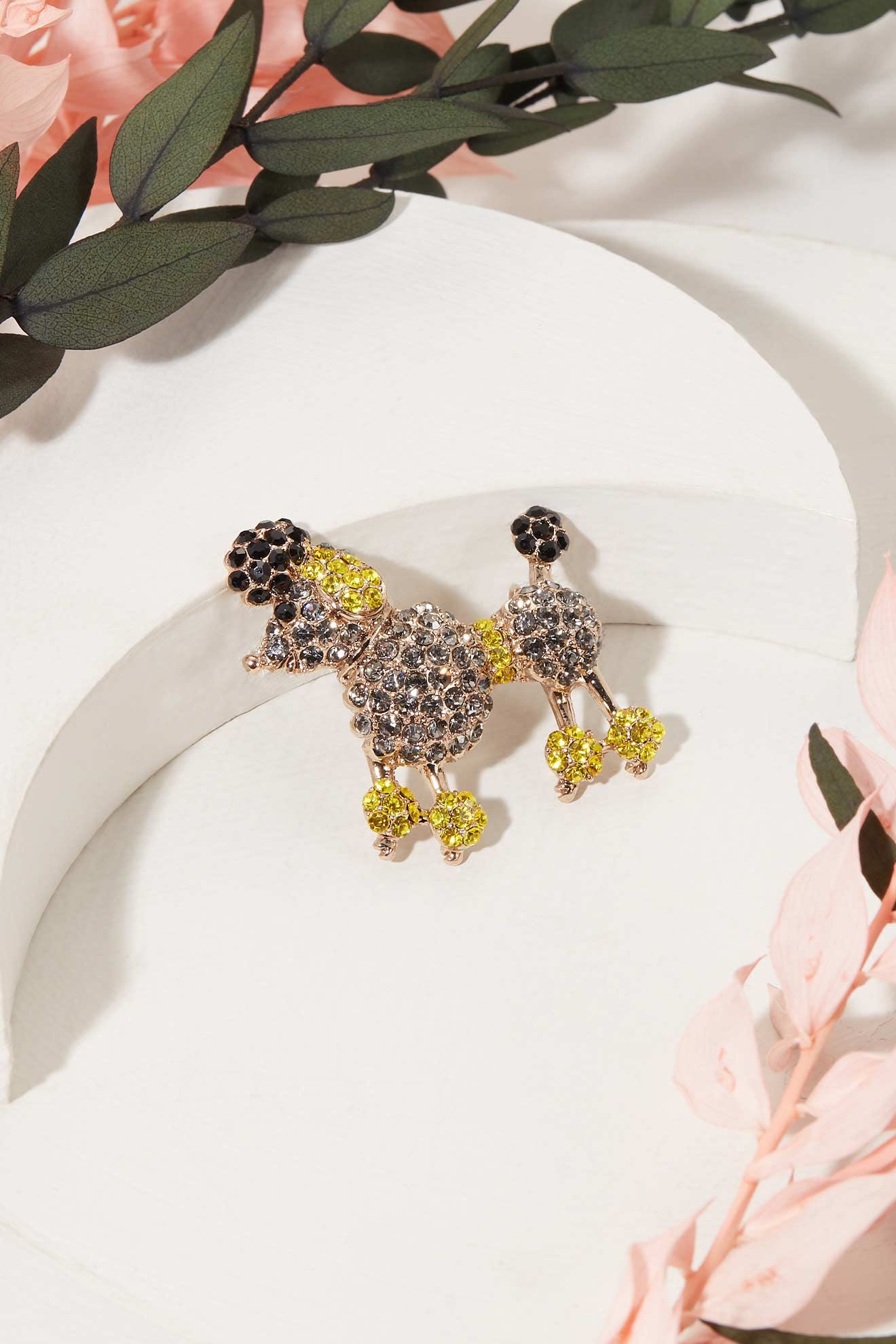 The Poodle Brooch is a whimsical gold accessory adorned with yellow and black rhinestones, shown on white with greenery and pink flowers.