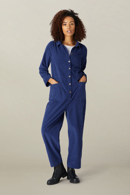 A woman with curly hair stands confidently wearing a dark blue long sleeve Cotton Babycord Jumpsuit and black boots against a neutral backdrop.