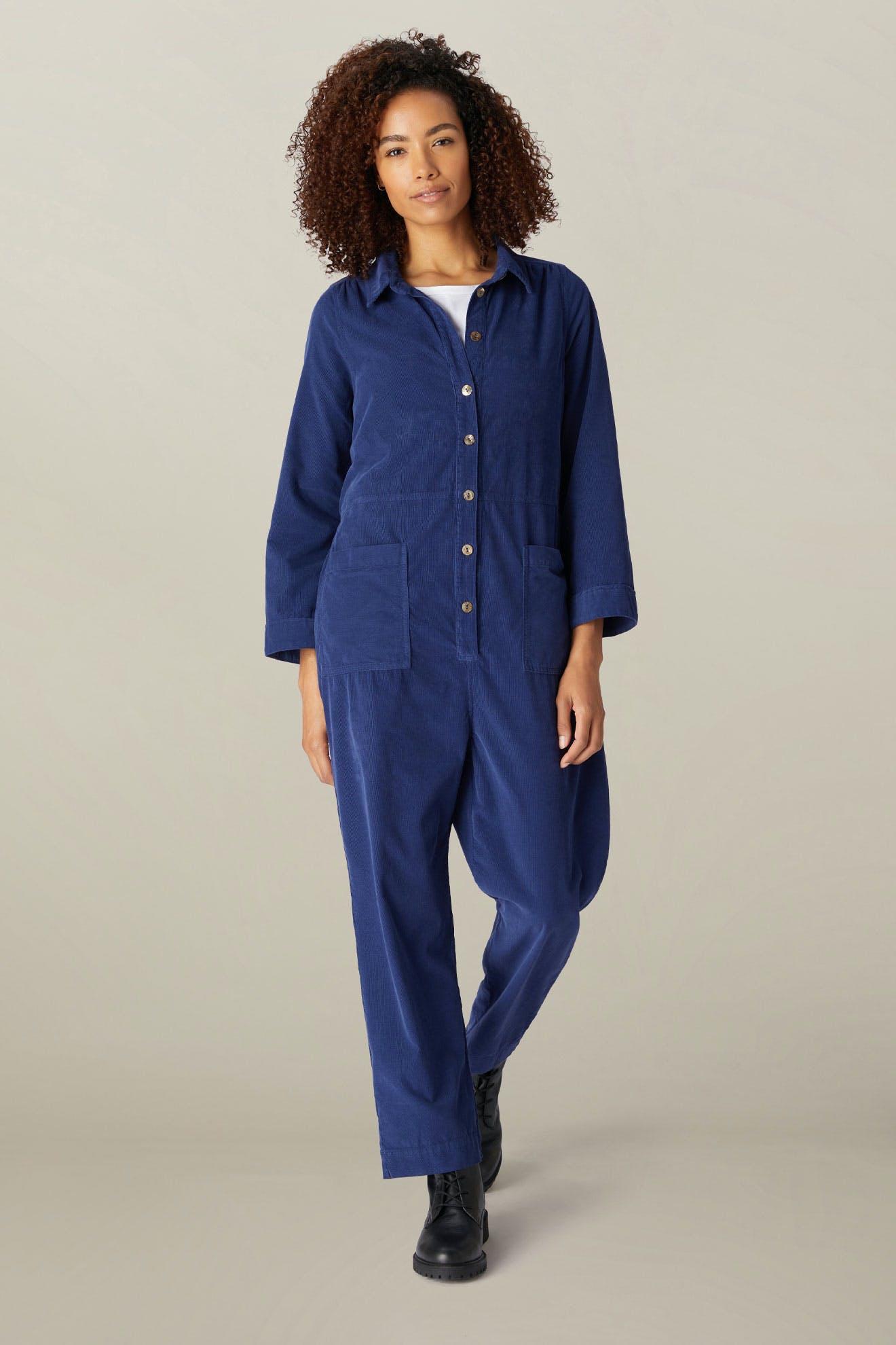 A woman in a blue Cotton Babycord Jumpsuit with cropped trouser legs standing confidently against a neutral background.