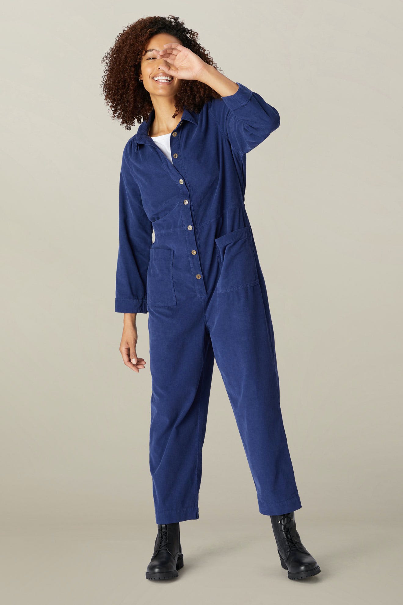 A woman in a blue Cotton Babycord jumpsuit and black boots smiling with one hand over her eyes, standing against a neutral backdrop.