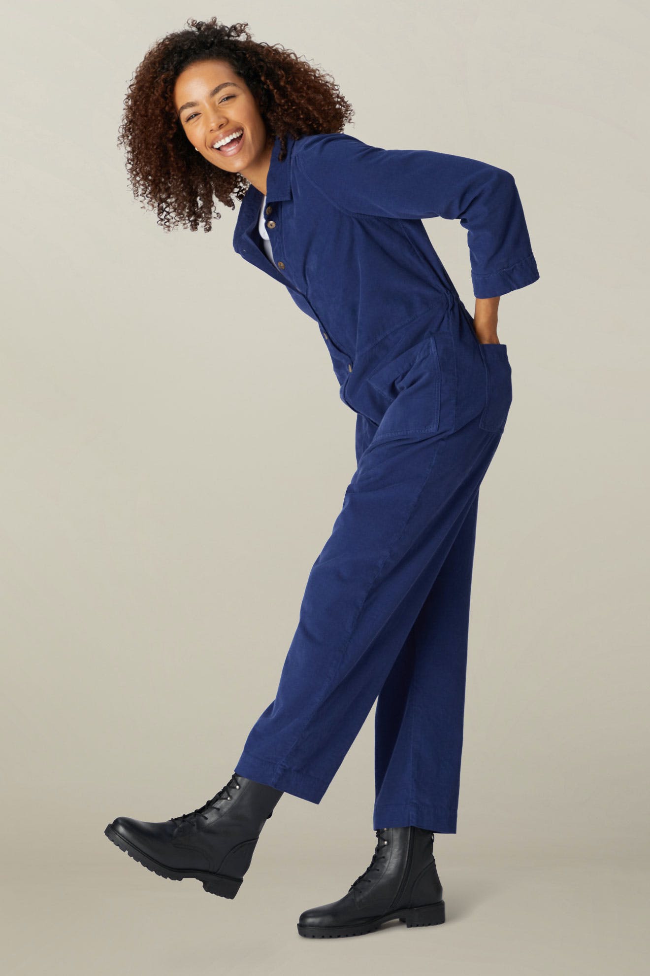 A woman in a Cotton Babycord Jumpsuit smiling and posing with one hand on her hip and wearing black boots, against a neutral background.