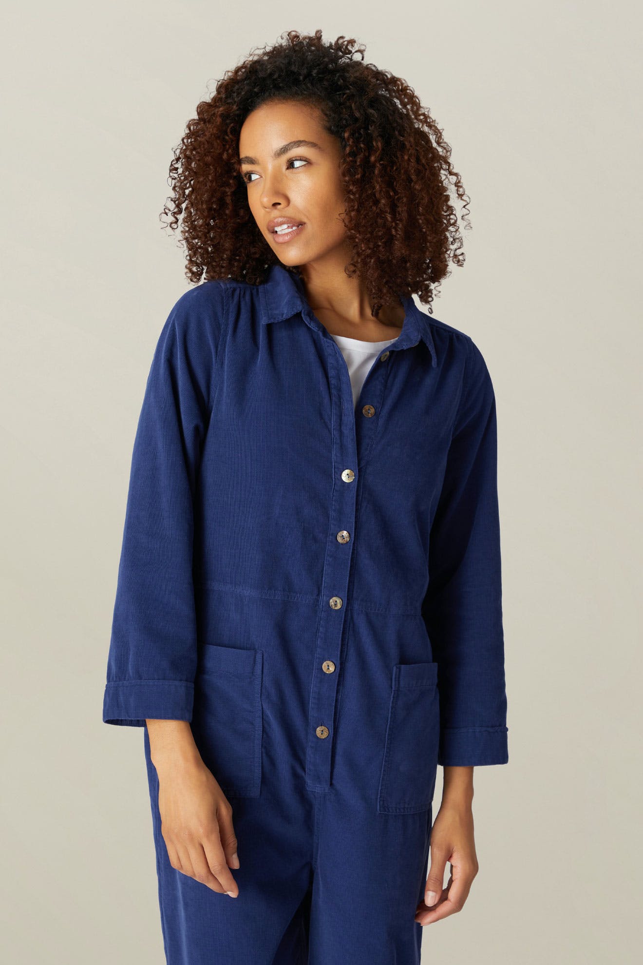 A woman with curly hair wearing a Cotton Babycord Jumpsuit in blue, looking to her right with a thoughtful expression.
