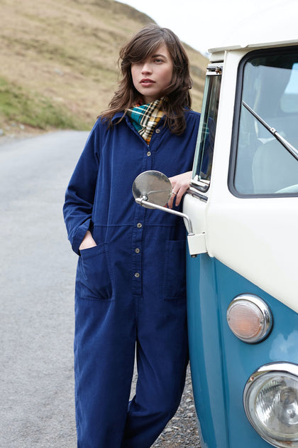 A woman in a blue Cotton Babycord jumpsuit and plaid scarf leaning on a vintage van, holding a mirror, with a grassy hill in the background.